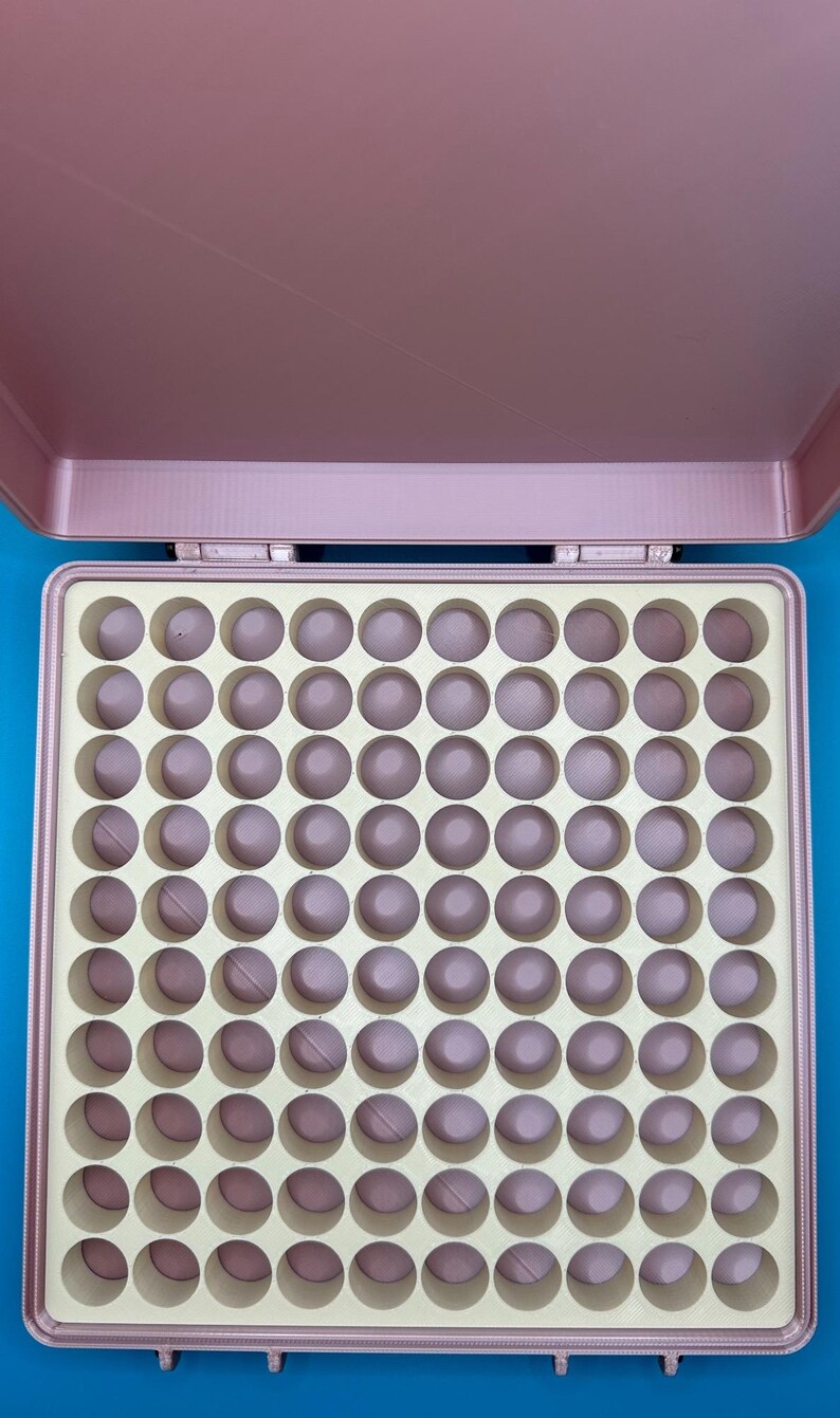 May include: A pink plastic storage case with a removable lid, revealing a beige foam insert with 100 round compartments.