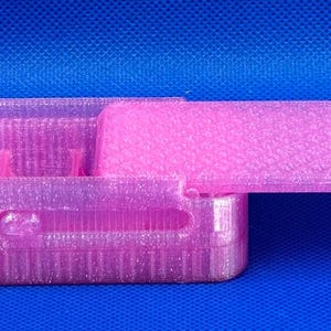 May include: A rectangular, translucent pink plastic storage box with a sliding lid. The box features a textured, glittery finish and internal dividers. The lid is partially open, revealing the hollow interior compartments of the container.