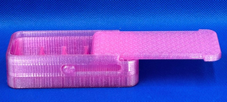 May include: A rectangular, translucent pink plastic storage box with a sliding lid. The box features a textured, glittery finish and internal dividers. The lid is partially open, revealing the hollow interior compartments of the container.