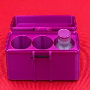 May include: A magenta rectangular storage case with an open hinged lid. The interior features three circular compartments, two of which are empty, while the third contains a small clear glass bottle with a matching magenta cap.