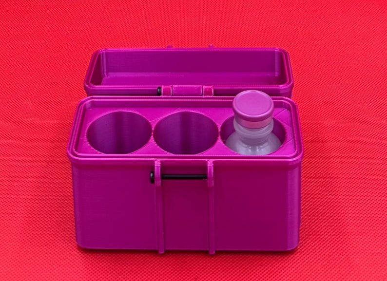 May include: A magenta rectangular storage case with an open hinged lid. The interior features three circular compartments, two of which are empty, while the third contains a small clear glass bottle with a matching magenta cap.
