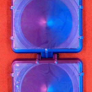 May include: A dual-hinged square device featuring a metallic purple and blue gradient finish. The surface displays a circular, concentric grooved pattern. The object is constructed with two identical square sections connected by a central hinge.