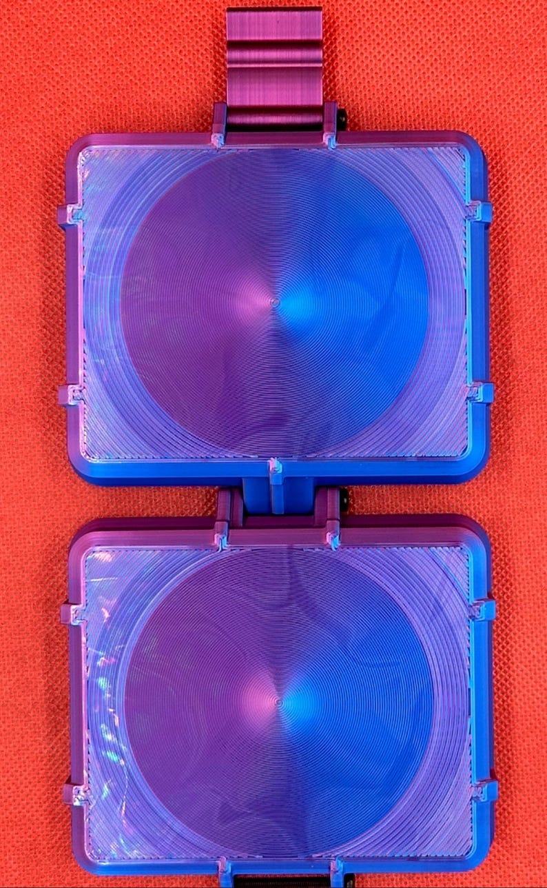 May include: A dual-hinged square device featuring a metallic purple and blue gradient finish. The surface displays a circular, concentric grooved pattern. The object is constructed with two identical square sections connected by a central hinge.