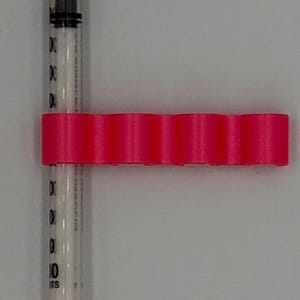 May include: A clear syringe with orange caps and a hot pink holder. The syringe has black markings for measurements. The holder has a series of rounded sections. The syringe is designed for medical use.