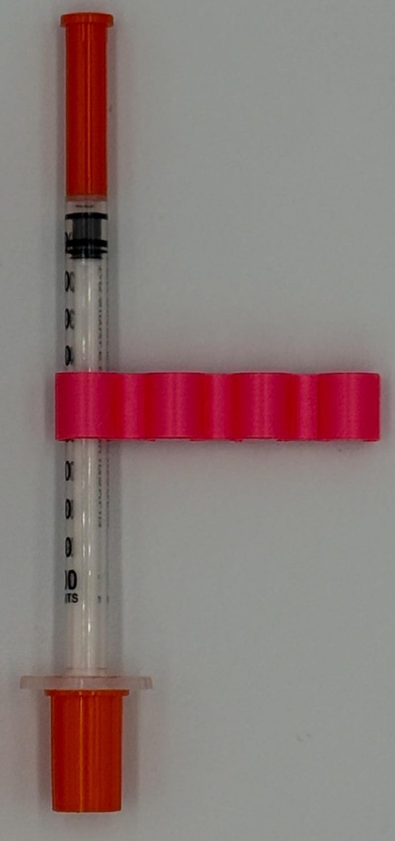 May include: A clear syringe with orange caps and a hot pink holder. The syringe has black markings for measurements. The holder has a series of rounded sections. The syringe is designed for medical use.