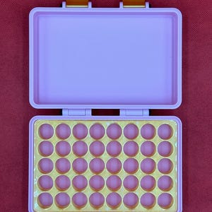 May include: Open, light purple case with a gold interior and pink circular indentations. The case has a rectangular shape with a hinged lid and gold-colored clasps. The background is a solid red color.