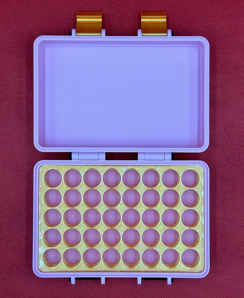 May include: Open, light purple case with a gold interior and pink circular indentations. The case has a rectangular shape with a hinged lid and gold-colored clasps. The background is a solid red color.