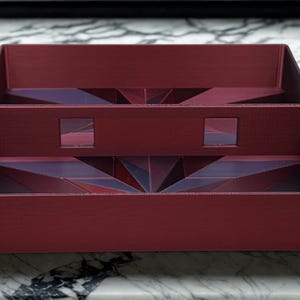 May include: A burgundy-colored, multi-compartment tray with geometric designs. The tray has a rectangular shape with interior dividers and cut-out squares. The geometric patterns are visible on the interior surfaces, adding visual interest to the design.