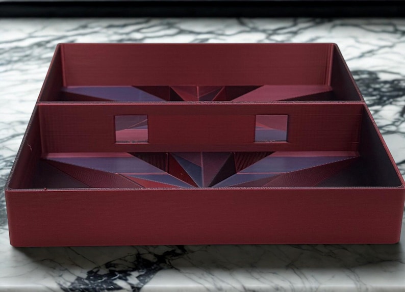 May include: A burgundy-colored, multi-compartment tray with geometric designs. The tray has a rectangular shape with interior dividers and cut-out squares. The geometric patterns are visible on the interior surfaces, adding visual interest to the design.