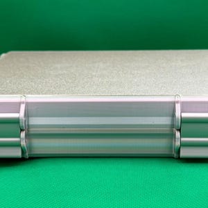 Puede incluir: Un estuche de almacenamiento rectangular de color plateado con una superficie superior texturizada y un marco met&aacute;lico liso. El estuche presenta dos bisagras plateadas prominentes en el borde frontal, aseguradas con peque&ntilde;os pasadores negros.