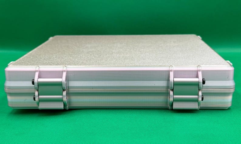 Puede incluir: Un estuche de almacenamiento rectangular de color plateado con una superficie superior texturizada y un marco met&aacute;lico liso. El estuche presenta dos bisagras plateadas prominentes en el borde frontal, aseguradas con peque&ntilde;os pasadores negros.