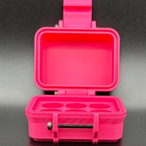 May include: A bright pink, rectangular storage case with an open lid. The interior base features six circular indentations for holding small items. A metal screw and a black threaded rod are visible on the front latch mechanism.