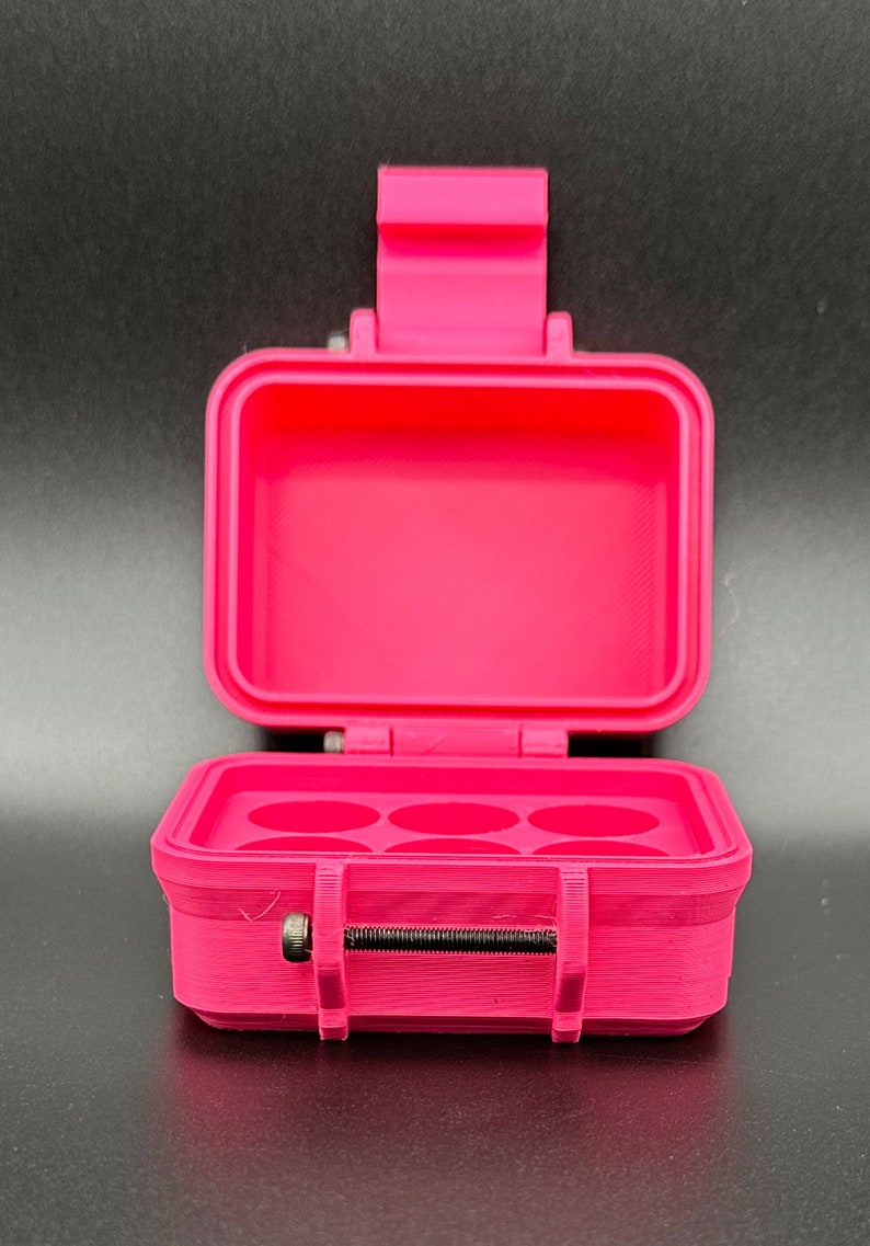 May include: A bright pink, rectangular storage case with an open lid. The interior base features six circular indentations for holding small items. A metal screw and a black threaded rod are visible on the front latch mechanism.