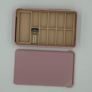 May include: A pink jewelry box with a divided interior and a separate lid. The interior has multiple compartments and a small glass vial. The box is open, revealing the compartments, and the lid is placed next to it.