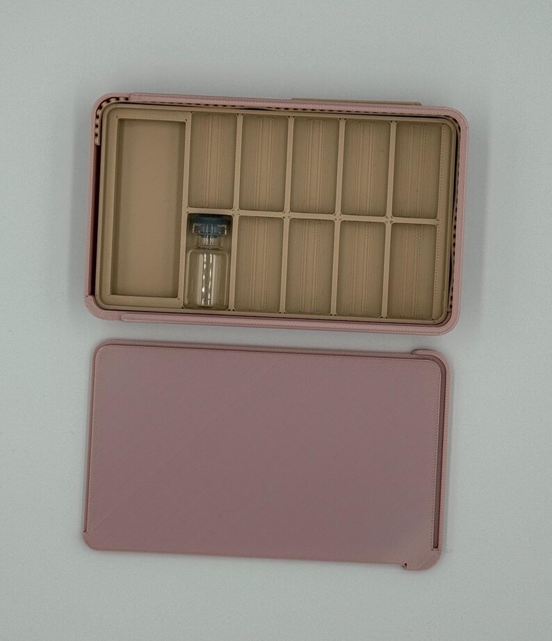 May include: A pink jewelry box with a divided interior and a separate lid. The interior has multiple compartments and a small glass vial. The box is open, revealing the compartments, and the lid is placed next to it.