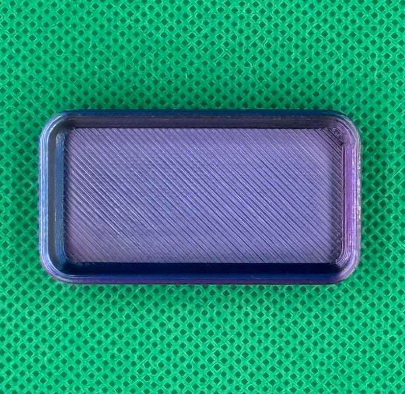 May include: A rectangular, 3D-printed tray with a textured, diagonal pattern in shades of purple and blue. The tray has rounded corners and a dark blue border, set against a green background.