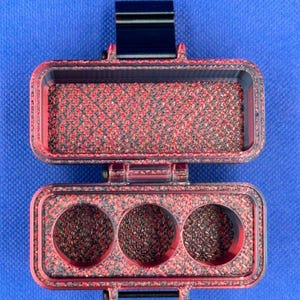 May include: A rectangular, hinged storage case with a red and black marbled texture. The top section is an open compartment, while the bottom section features three circular cutouts. A black latch is visible at the top and a hinge at the bottom.