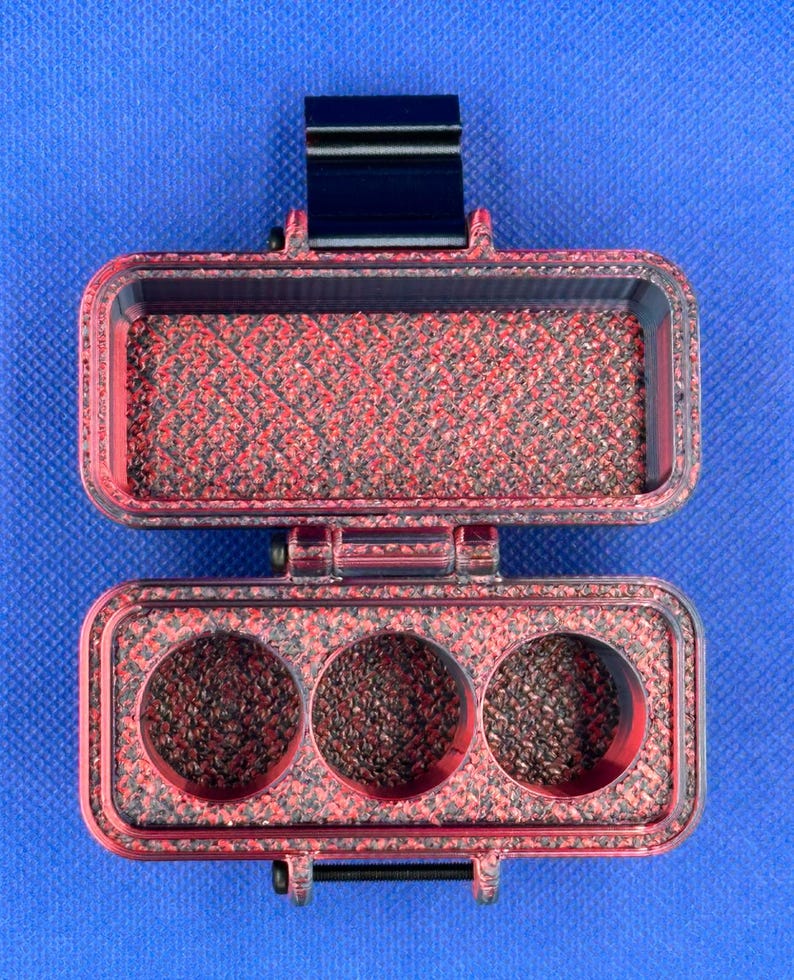 May include: A rectangular, hinged storage case with a red and black marbled texture. The top section is an open compartment, while the bottom section features three circular cutouts. A black latch is visible at the top and a hinge at the bottom.