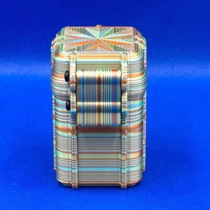 May include: A rectangular, multi-colored storage container featuring a layered, striped pattern in shades of orange, teal, and cream. The box has a hinged lid with a central starburst design and two small black circular fasteners on the side.