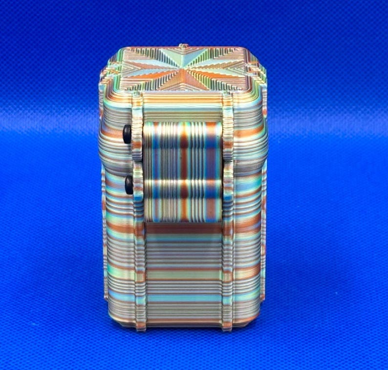 May include: A rectangular, multi-colored storage container featuring a layered, striped pattern in shades of orange, teal, and cream. The box has a hinged lid with a central starburst design and two small black circular fasteners on the side.