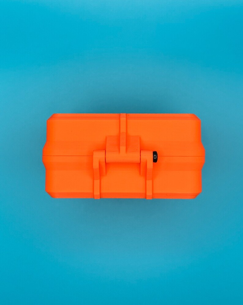 May include: A bright orange rectangular hard-shell storage case featuring a central latch mechanism and a small black circular pin detail on the side, presented in a closed position with a clean, uniform exterior surface design.