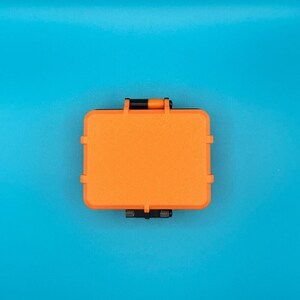 May include: A rectangular, bright orange protective storage case with a textured surface. The case features black hinges and a black latch mechanism on the top and bottom edges, designed for secure closure and durability in various environments.