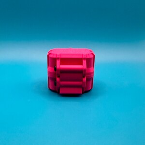 May include: A bright pink, rectangular, hard-shell storage case with a textured surface. The case features a prominent central latch mechanism with two small black circular accents on the side, designed for secure closure and protection.