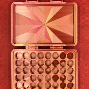 May include: A rectangular storage case with a hinged lid, featuring a metallic copper and pink gradient finish. The interior lid displays a geometric starburst pattern, while the base contains a grid of thirty-five circular compartments.