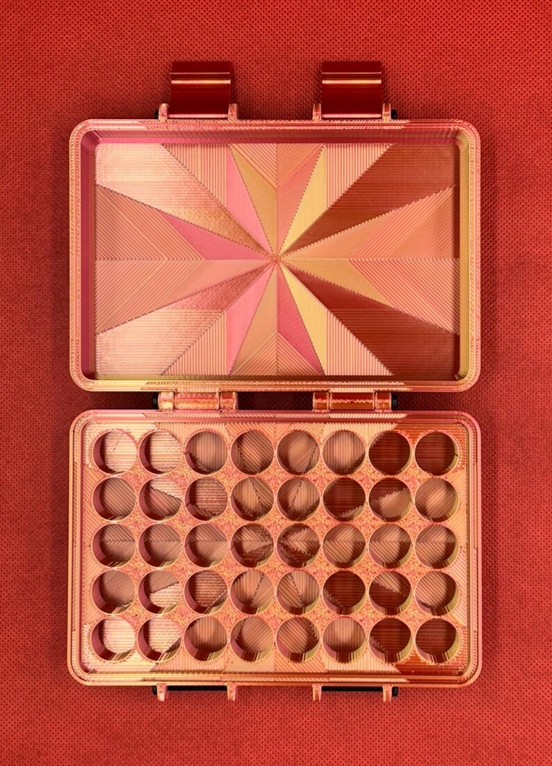 May include: A rectangular storage case with a hinged lid, featuring a metallic copper and pink gradient finish. The interior lid displays a geometric starburst pattern, while the base contains a grid of thirty-five circular compartments.
