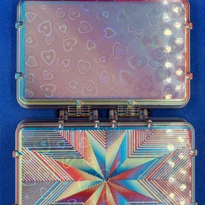 May include: A rectangular, iridescent storage case with a hinged lid, featuring a holographic heart pattern and a central starburst design. Two plaid fabric loops are attached to the top edge of the case, which displays a metallic, multi-colored finish.