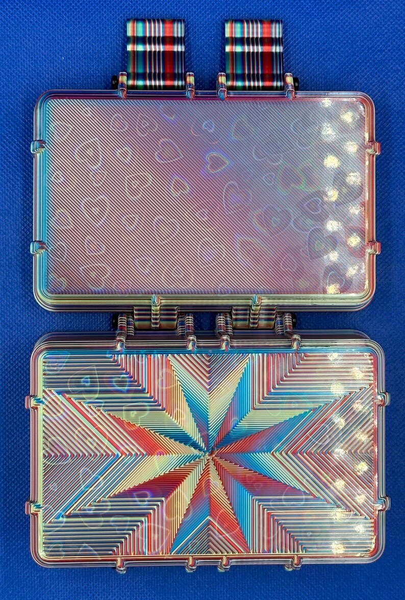 May include: A rectangular, iridescent storage case with a hinged lid, featuring a holographic heart pattern and a central starburst design. Two plaid fabric loops are attached to the top edge of the case, which displays a metallic, multi-colored finish.