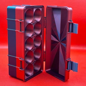 Puede incluir: Una caja de almacenamiento rectangular con tapa abatible, con un exterior rojo y azul. El interior contiene diez compartimentos circulares dispuestos en dos columnas, con un patr&oacute;n geom&eacute;trico de estrella en la tapa.