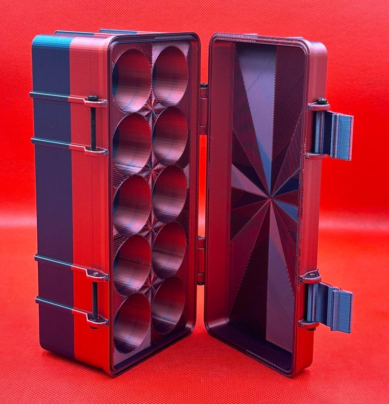 Puede incluir: Una caja de almacenamiento rectangular con tapa abatible, con un exterior rojo y azul. El interior contiene diez compartimentos circulares dispuestos en dos columnas, con un patr&oacute;n geom&eacute;trico de estrella en la tapa.