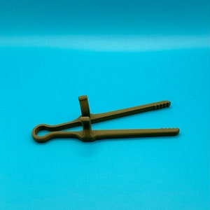 May include: A matte olive green plastic kitchen tong featuring a circular handle loop at one end, two long arms with serrated tips, and two perpendicular upright tabs located near the center hinge point for gripping and control.