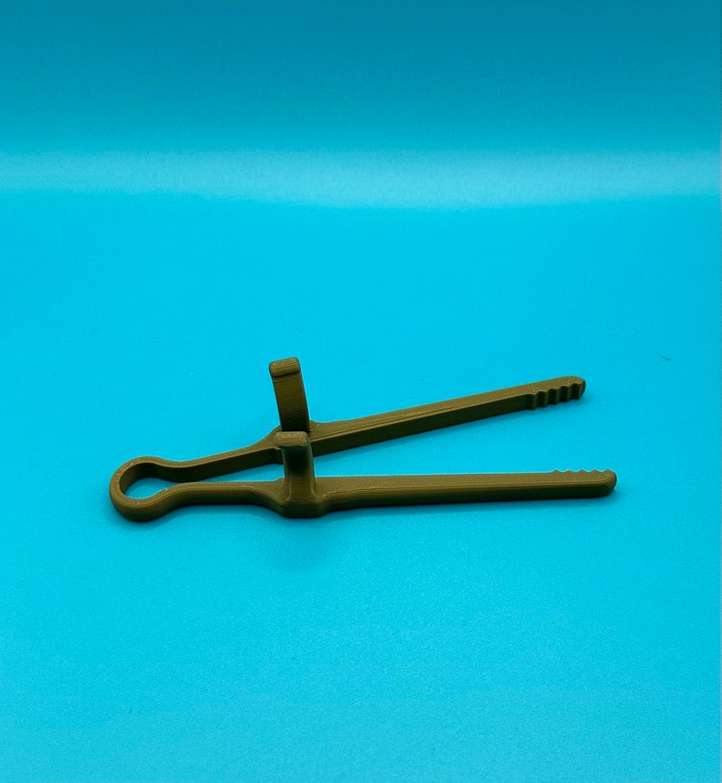 May include: A matte olive green plastic kitchen tong featuring a circular handle loop at one end, two long arms with serrated tips, and two perpendicular upright tabs located near the center hinge point for gripping and control.