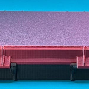 May include: A rectangular storage case featuring a black base and a dark red lid with a textured, glittery purple top surface. The case has two visible hinges connecting the lid to the base, with a structured, layered design on the sides.