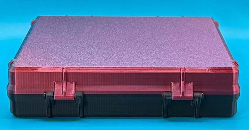 May include: A rectangular storage case featuring a black base and a dark red lid with a textured, glittery purple top surface. The case has two visible hinges connecting the lid to the base, with a structured, layered design on the sides.