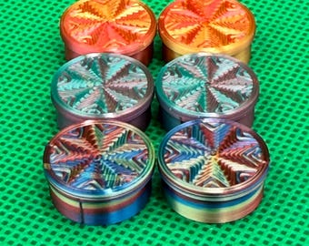 Op de afbeelding: Zes ronde metalen grinders gerangschikt in twee kolommen. De bovenste twee hebben een oranje en gele afwerking, de middelste twee hebben een blauwgroen en paars verloop, en de onderste twee tonen een meerkleurig iriserend patroon.