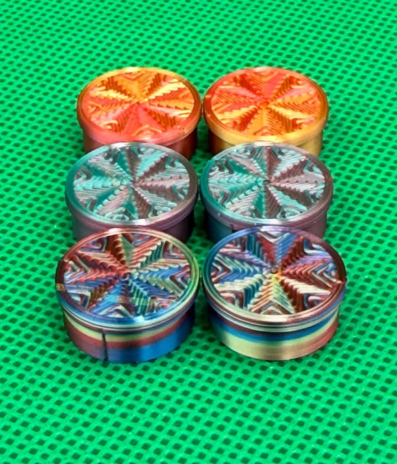 May include: Six circular metal grinders arranged in two columns. The top two have an orange and yellow finish, the middle two feature a teal and purple gradient, and the bottom two display a multi-colored iridescent pattern with star-like textures.