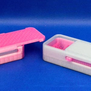 May include: Two rectangular storage boxes with sliding lids. The left box is bright pink with a textured lid and a small grey latch. The right box is translucent white with a smooth lid and a pink interior compartment visible inside.