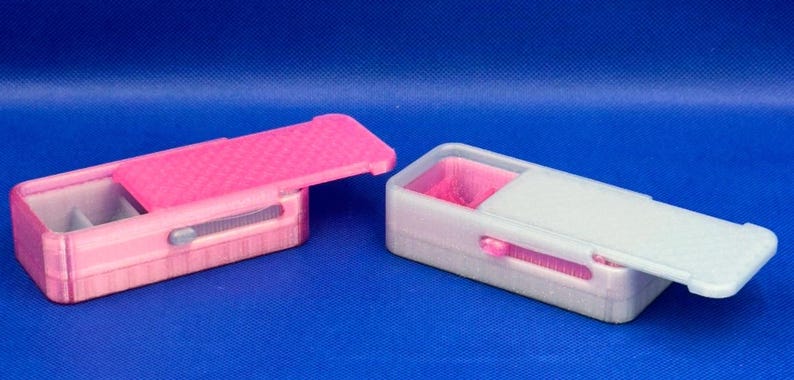 May include: Two rectangular storage boxes with sliding lids. The left box is bright pink with a textured lid and a small grey latch. The right box is translucent white with a smooth lid and a pink interior compartment visible inside.