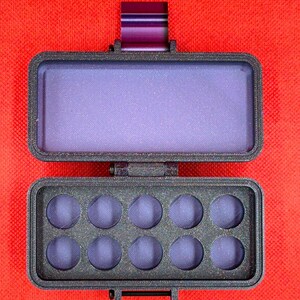 May include: A rectangular, dark gray, textured plastic palette case with a hinged lid. The interior features ten circular recessed wells for paint, and the lid contains a smooth, flat purple surface. A purple metal latch is visible at the top.