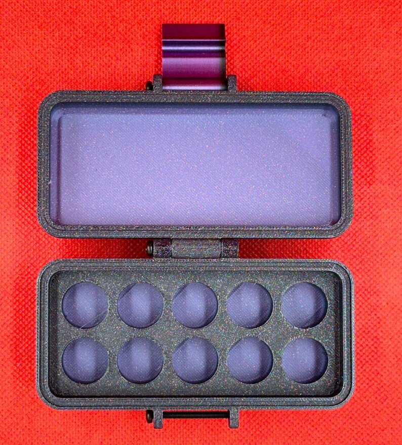 May include: A rectangular, dark gray, textured plastic palette case with a hinged lid. The interior features ten circular recessed wells for paint, and the lid contains a smooth, flat purple surface. A purple metal latch is visible at the top.