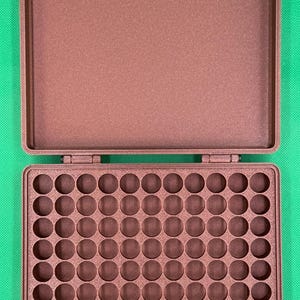 K&ouml;nnte beinhalten: Ein brauner, rechteckiger Aufbewahrungskoffer mit Klappdeckel. Der Innenboden verf&uuml;gt &uuml;ber ein Raster aus einhundert kreisf&ouml;rmigen F&auml;chern, die in zehn Reihen und zehn Spalten angeordnet sind, um kleine, runde Gegenst&auml;nde strukturiert aufzubewahren.