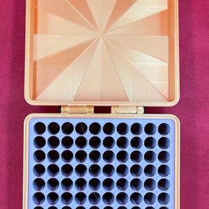 May include: A square, peach-colored storage case with a hinged lid. The interior of the lid features a sunburst pattern. The base contains a light purple insert with a grid of one hundred circular holes for organizing small items.