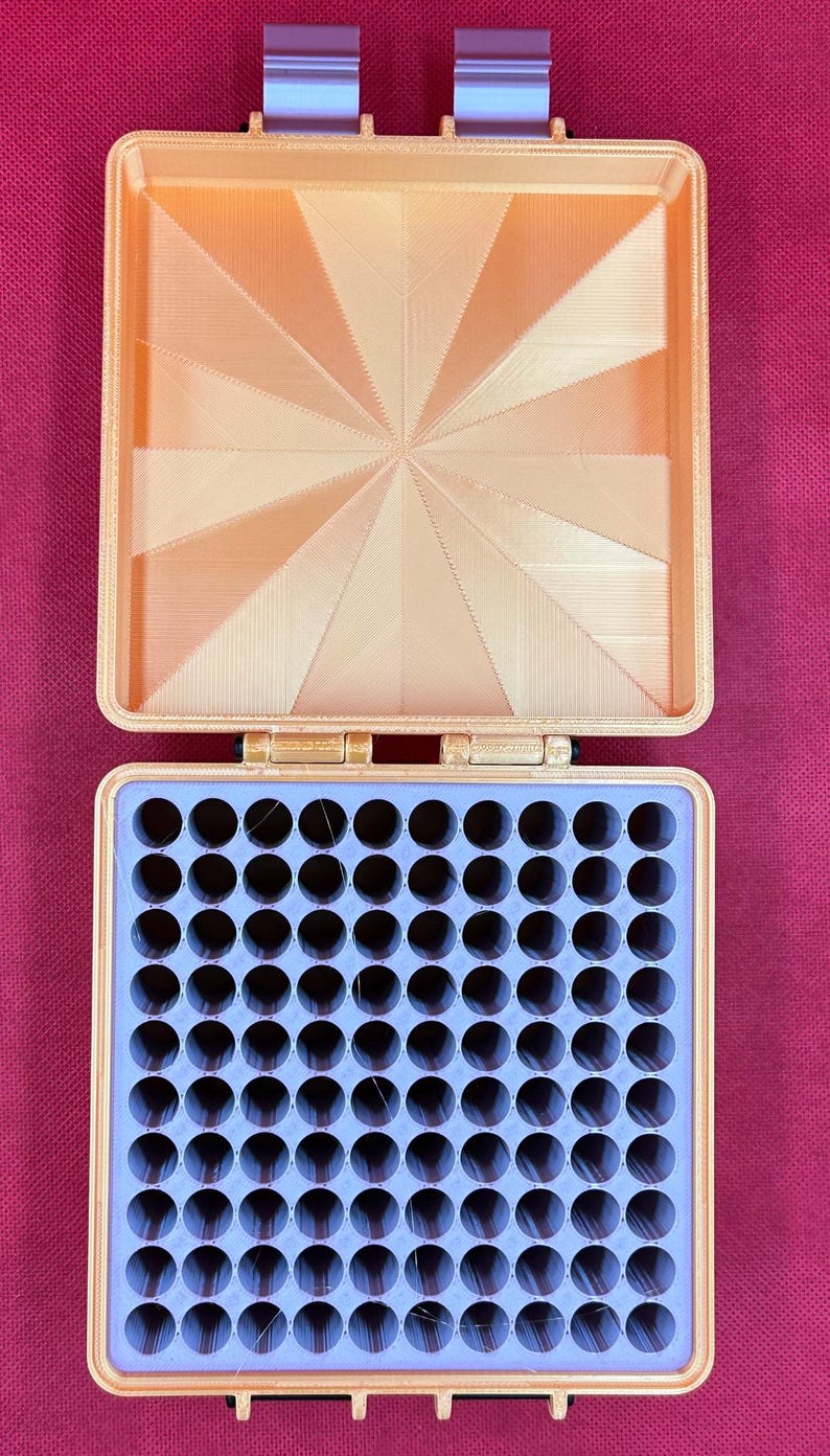 May include: A square, peach-colored storage case with a hinged lid. The interior of the lid features a sunburst pattern. The base contains a light purple insert with a grid of one hundred circular holes for organizing small items.