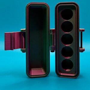 Puede incluir: Un estuche de almacenamiento rectangular impreso en 3D con tapa abatible, que presenta un acabado iridiscente que cambia de color entre violeta, verde y negro. El interior contiene cinco compartimentos circulares para guardar objetos.