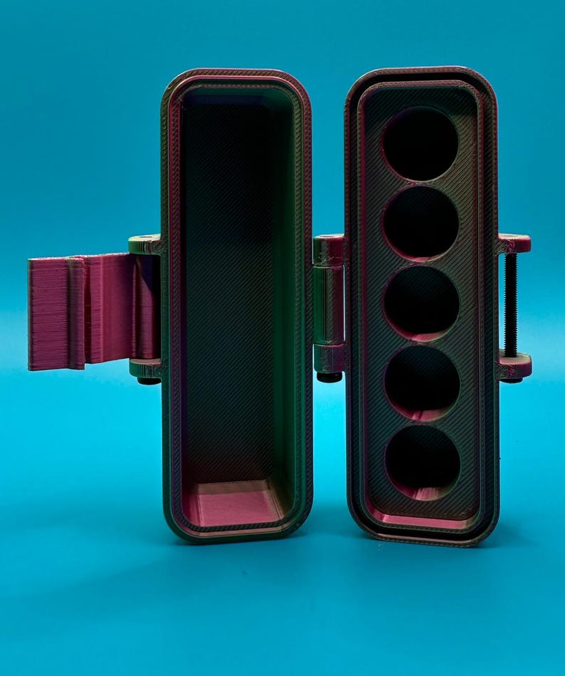 Puede incluir: Un estuche de almacenamiento rectangular impreso en 3D con tapa abatible, que presenta un acabado iridiscente que cambia de color entre violeta, verde y negro. El interior contiene cinco compartimentos circulares para guardar objetos.