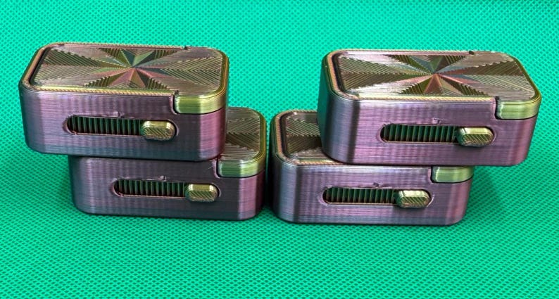 May include: Four rectangular, iridescent purple and gold fidget sliders stacked in two pairs. Each slider features a textured, starburst-patterned top surface and a horizontal side slot containing a sliding gold-coloured metal toggle mechanism.