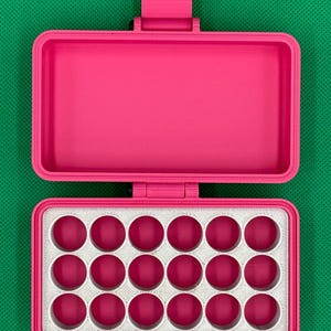 K&ouml;nnte beinhalten: Ein leuchtend pinkfarbener, rechteckiger Aufbewahrungskoffer mit Klappdeckel. Das Innere verf&uuml;gt &uuml;ber einen wei&szlig;en Einsatz mit achtzehn kreisf&ouml;rmigen Aussparungen in drei Reihen zu je sechs, die den pinken Boden durch jede &Ouml;ffnung zeigen.